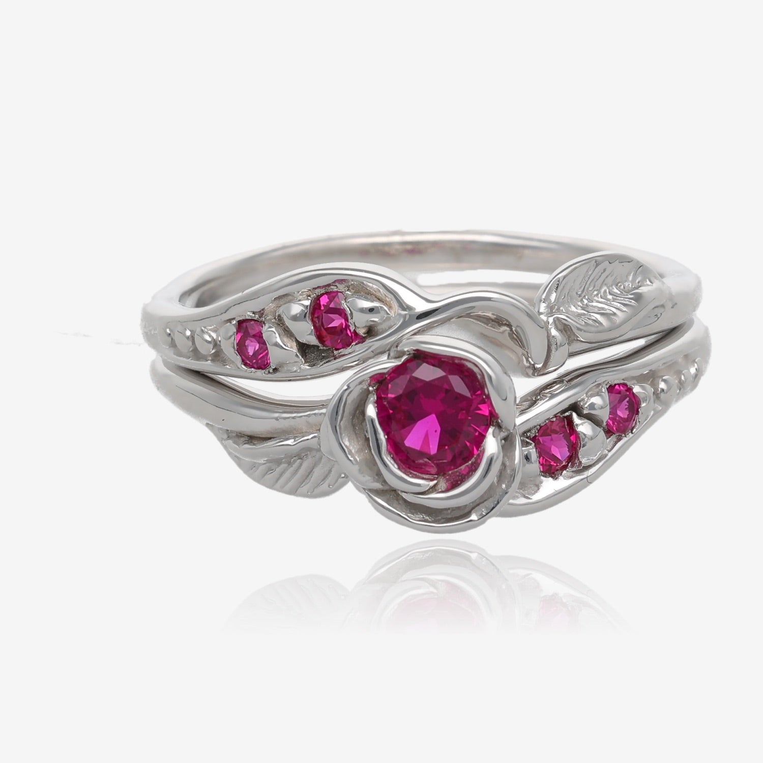 Photo shows a silver wedding set with a rosebud center. A single round pinkish red ruby is set into the petals of the rose. A leaf is placed on each side of the bloom, facing outward diagonally. Smaller deep pink gems are set along the sides