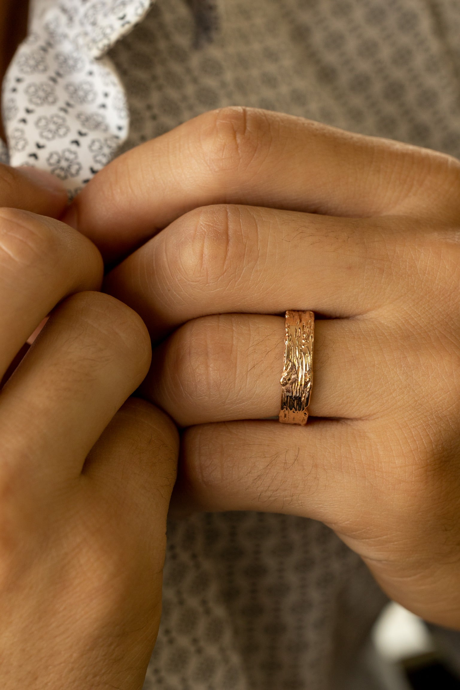 Image shows man’s hand wearing a gold band approximately one quarter inch wide, textured like a slice of birch tree. The ring profile is flat, with the raised and recessed bark texture running across the finger.