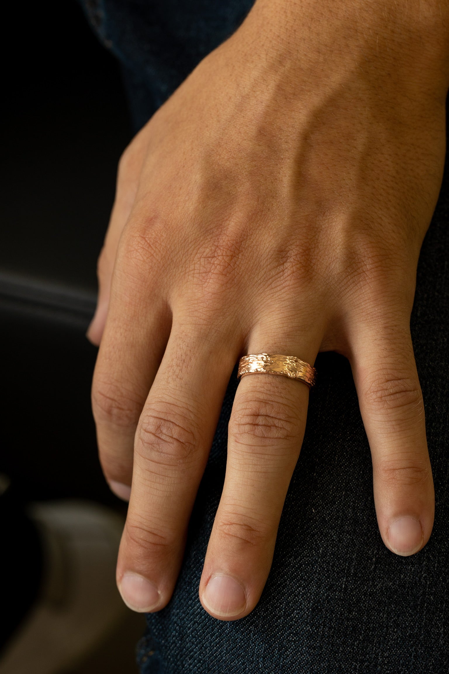 Image shows man’s hand wearing a gold band approximately one quarter inch wide, textured like a slice of birch tree. The ring profile is flat, with the raised and recessed bark texture running across the finger.
