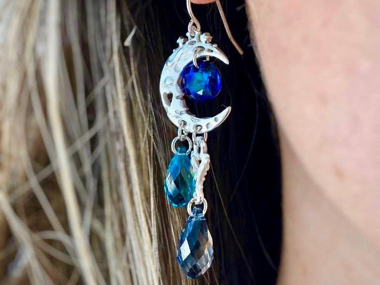 Front view of a silver earring shaped like a crescent moon with a dark blue circle-shaped gem bead in the center of the crescent, a heart shaped charm on the bottom of the earring located towards the point of the crescent, and 2 light blue tear-shaped gem beads on the bottom of the earring. The earring is on a white, brunette model.