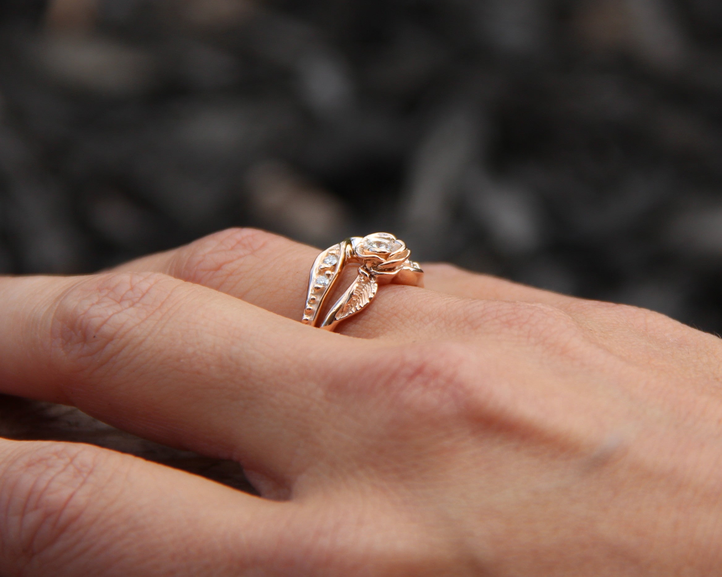 woman’s hand wears a rose bloom on a rose gold wedding ring set that measures half an inch wide. The rose is 0.25 inch across and holds a round diamond, and the bands curve to fit together. Small leaves and diamonds offset each side