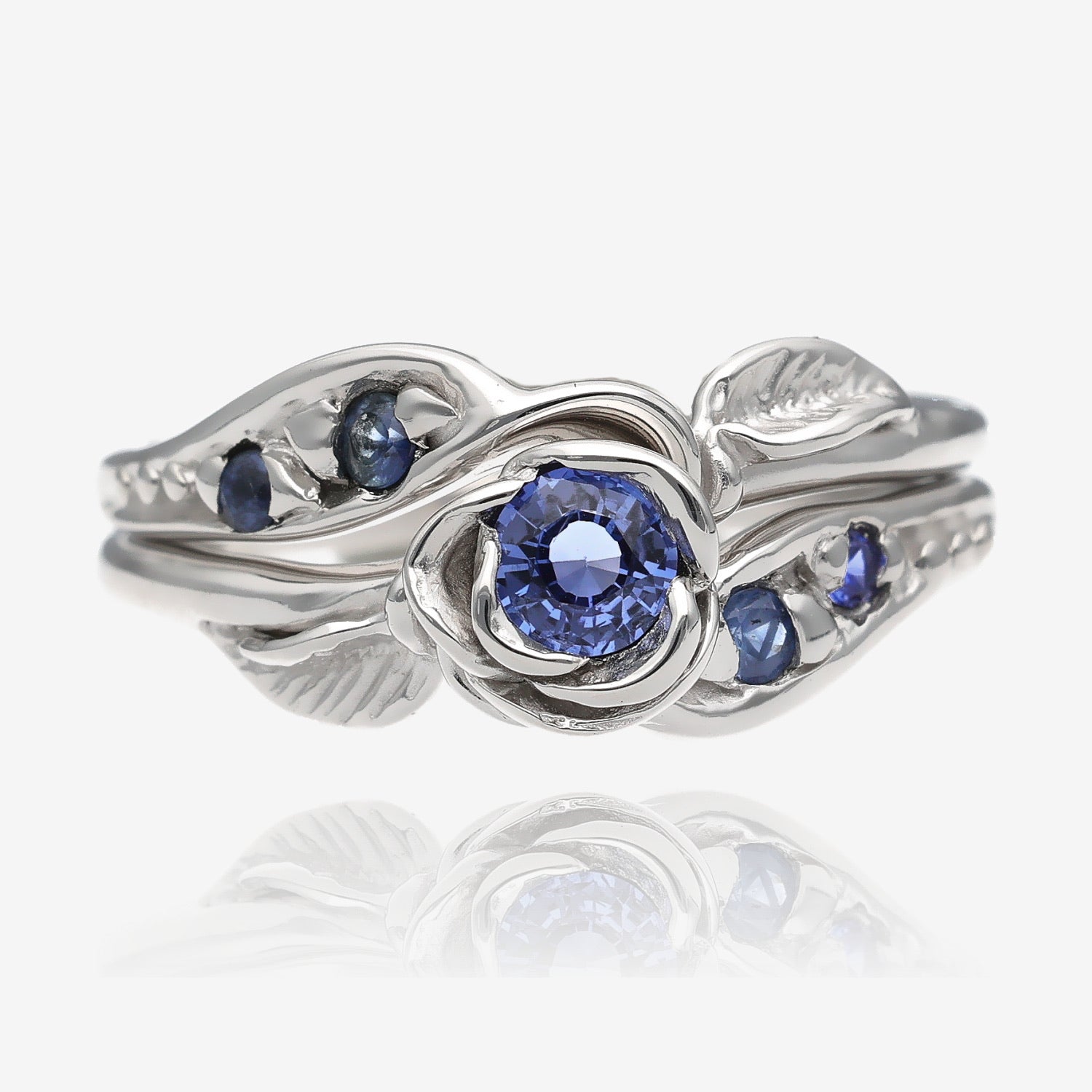 Photo shows a silver wedding set with a rosebud center. A single round lavender blue tanzanite is set into the petals of the rose. A leaf is placed on each side of the bloom, facing outward diagonally. Four more tanzanite gems are set on the sides