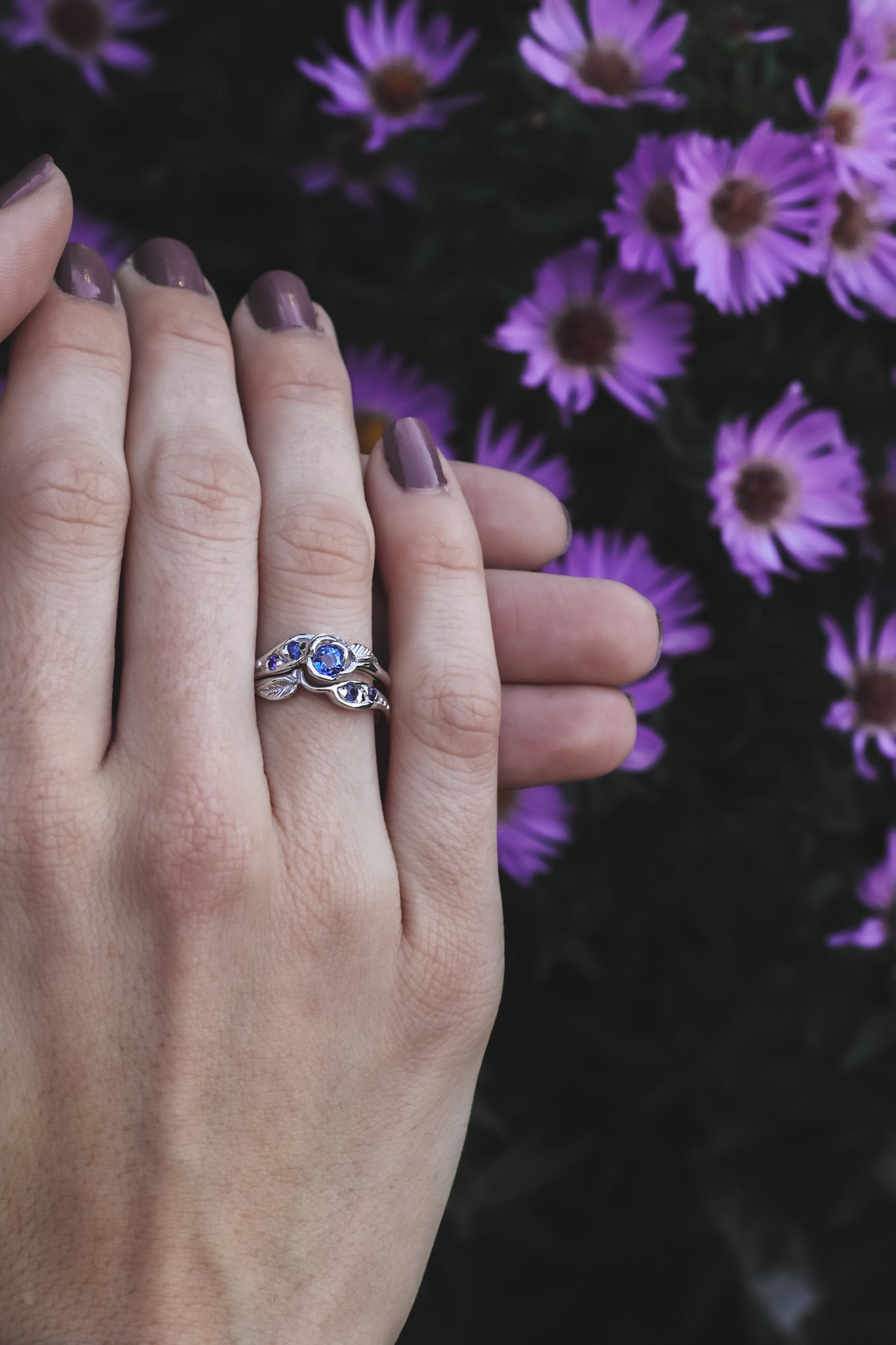 Woman’s hand wearing a ring set with a rosebud center. A single round lavender blue tanzanite is set into the petals of the rose. A leaf is placed on each side of the bloom, facing outward diagonally. Four more tanzanite gems are set on the sides