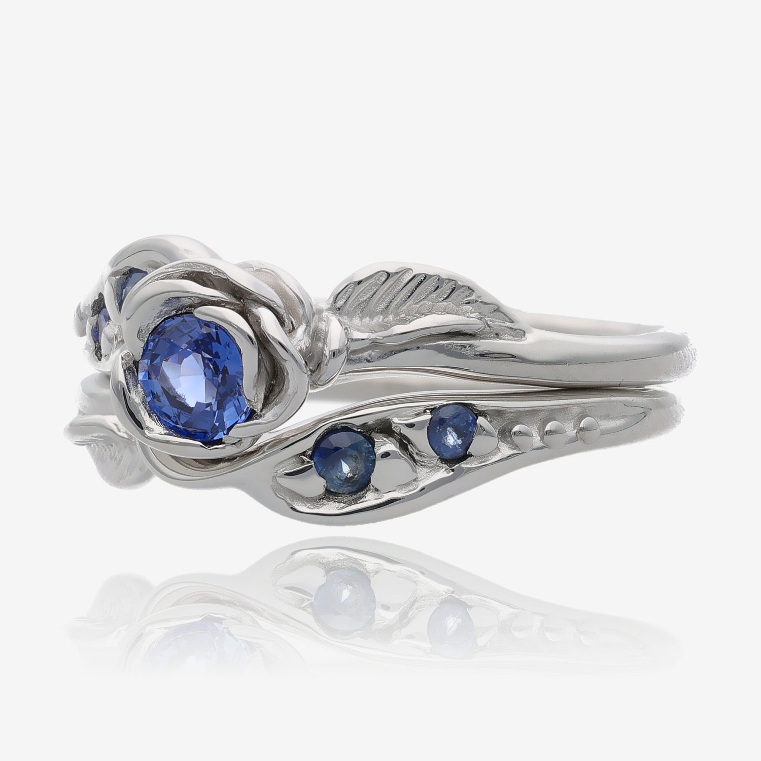 Side view of silver wedding set with a rosebud center. round lavender blue tanzanite is set into the petals of the rose. A leaf is placed on each side of the bloom, curving down the band. Accent tanzanite gems are set on the sides