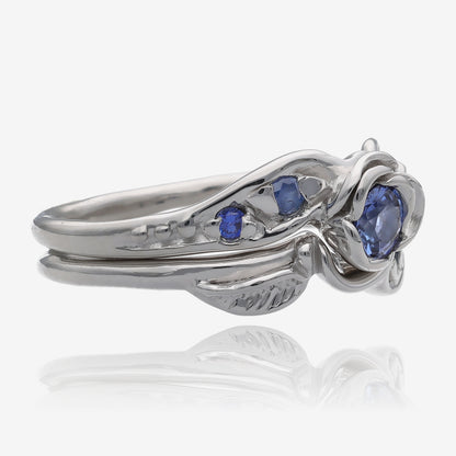 Side view of silver wedding set with a rosebud center. round lavender blue tanzanite is set into the petals of the rose. A leaf is placed on each side of the bloom, curving down the band. Accent tanzanite gems are set on the sides