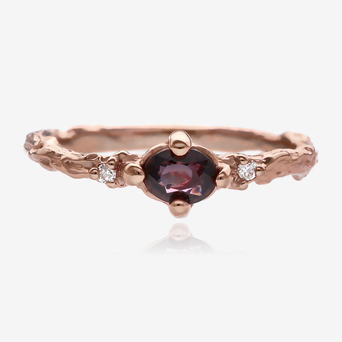 Plum purple faceted oval sapphire is set lengthwise in a slender, twig textured rose gold band with a small diamond on each side