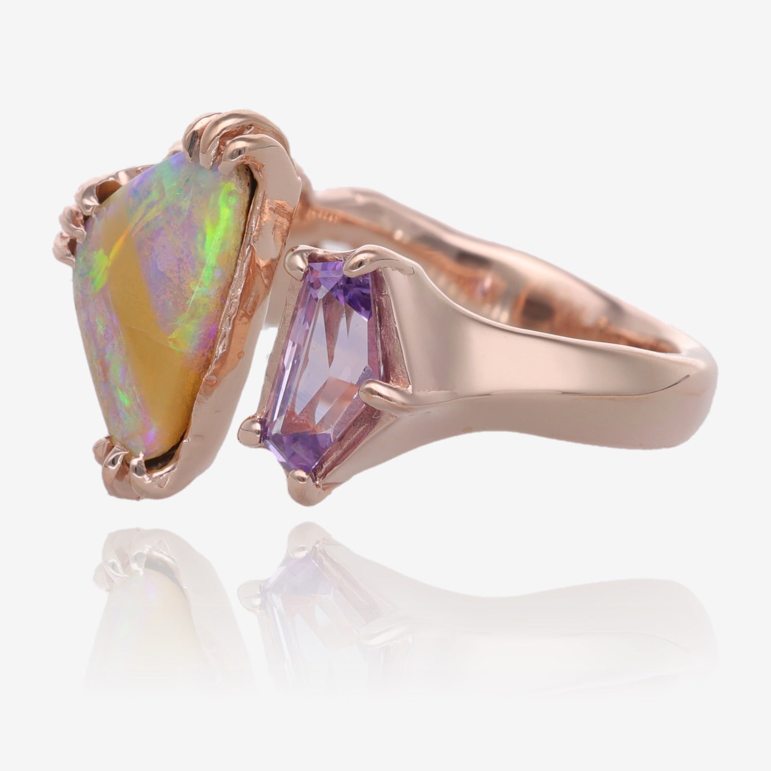 Side view of Rose gold ring with opal on left side. Opal is triangular in shape and has a lavender and green pattern. Each corner of the triangle has three thin prong settings. There is a gap on top of the ring which is part of the design. The other gem is a pale lavender five sided sapphire with a prong at each corner. 
Ring band is 3.5mm on bottom. 