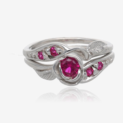 Photo shows a silver wedding set with a rosebud center. A single round pinkish red ruby is set into the petals of the rose. A leaf is placed on each side of the bloom, facing outward diagonally. Smaller deep pink gems are set along the sides