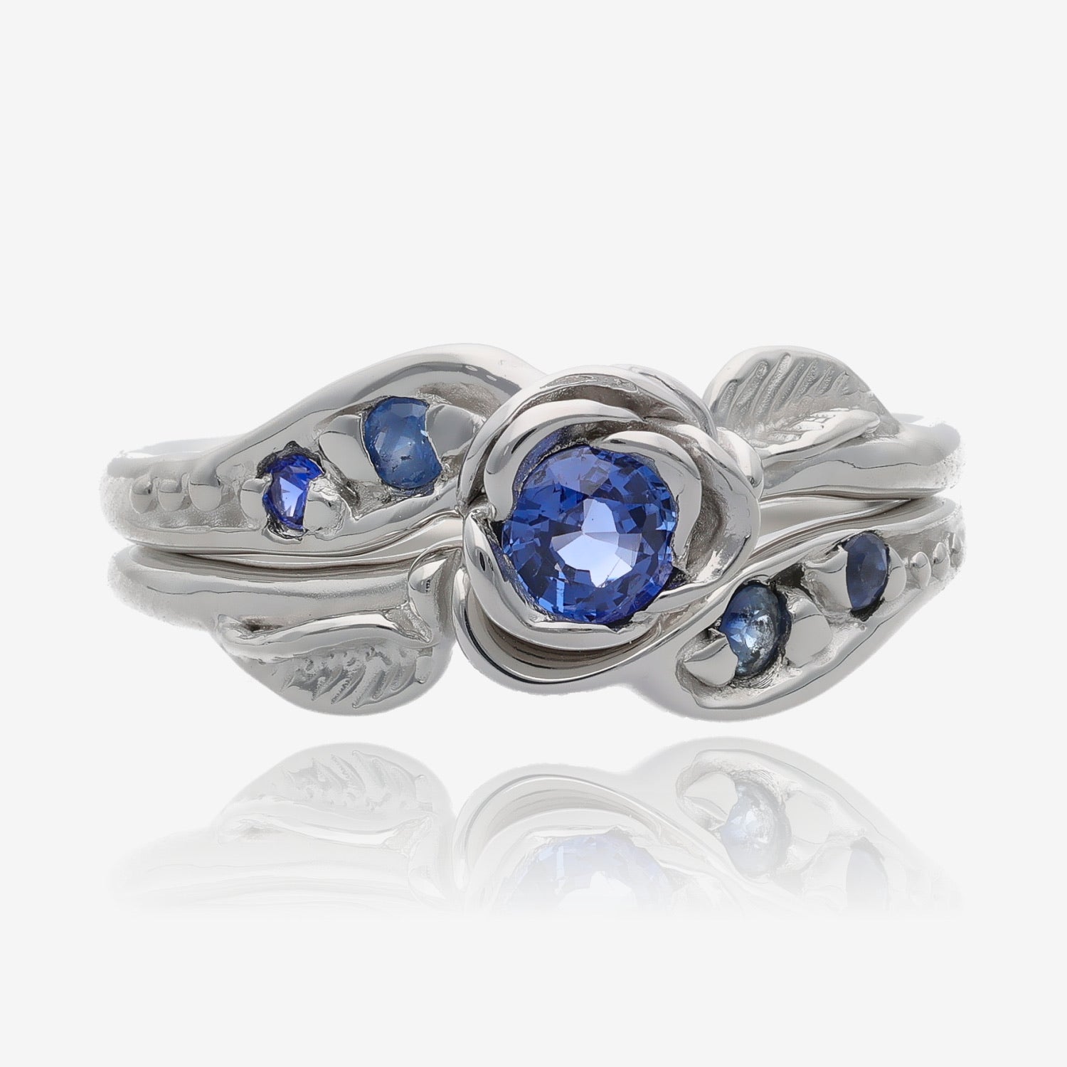 Photo shows a silver wedding set with a rosebud center. A single round lavender blue tanzanite is set into the petals of the rose. A leaf is placed on each side of the bloom, facing outward diagonally. Four more tanzanite gems are set on the sides