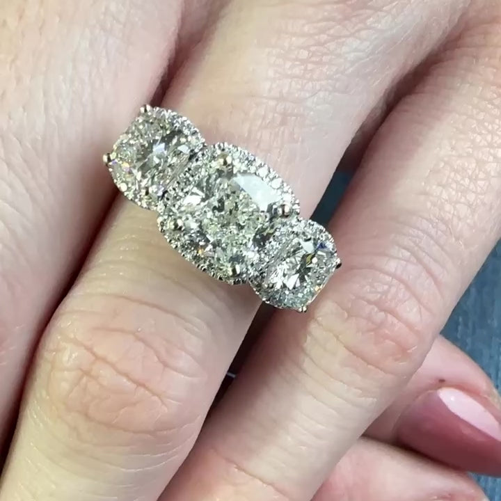 Woman’s hand wears a white gold ring with three large cushion shaped diamonds, each surrounded by a halo of small diamonds. All stones set in prongs