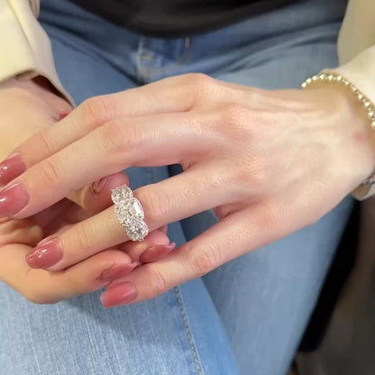 Woman’s hand wears a white gold ring with three large cushion shaped diamonds, each surrounded by a halo of small diamonds. All stones set in prongs