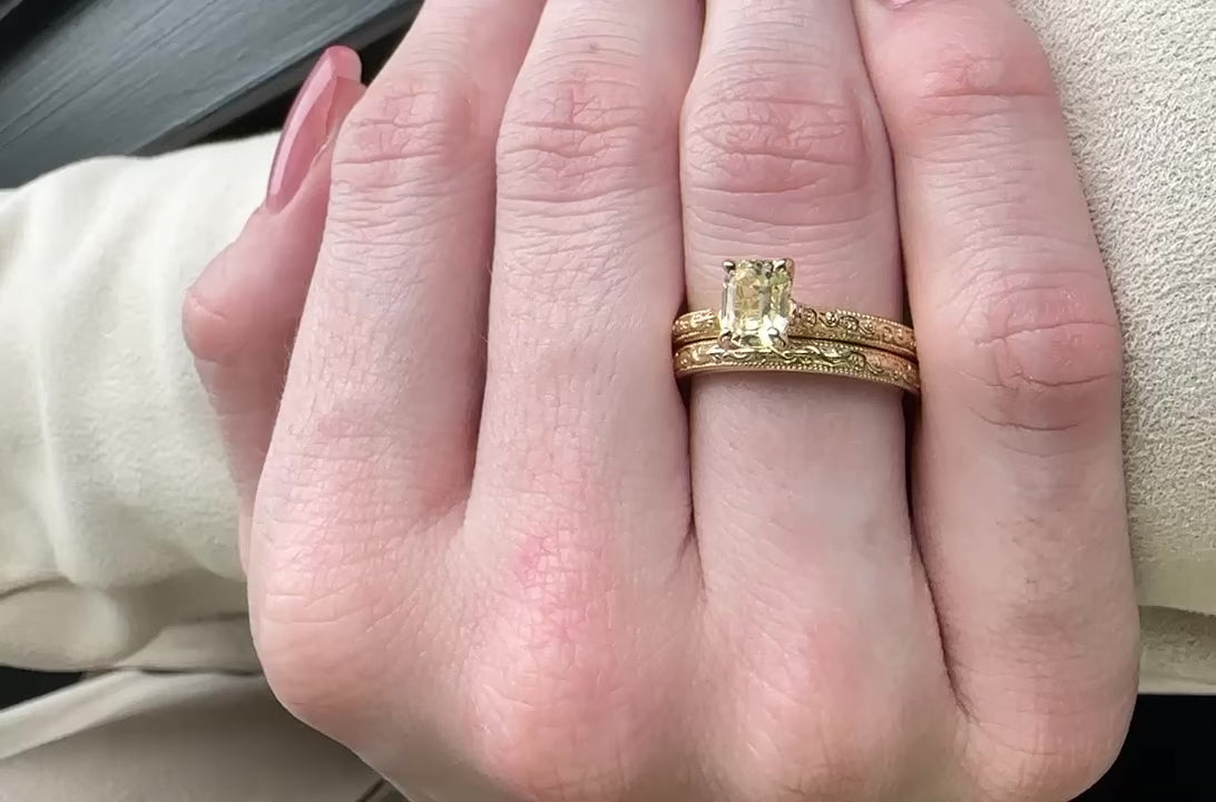 A woman’s hand wears this yellow gold wedding set. Ring set features a solitaire, light, yellow gemstone in a rectangular step cut facet style.