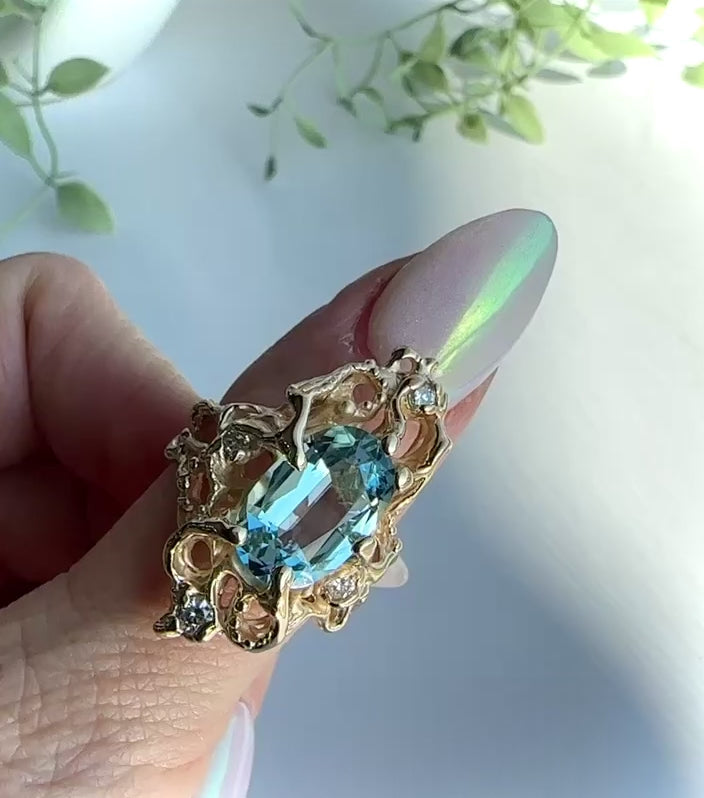 Gold and aquamarine ring is shown on a woman’s thumb. In the video the hand rotates to show the design undulating and curling around the finger. The woman’s thumbnail is long and pearl colored, background is light gray with some greenery 