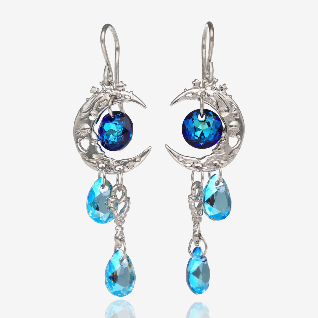 Front view of 2 silver earrings shaped like a crescent moon with a dark blue circle-shaped gem bead in the center of the crescent, a heart shaped charm on the bottom of the earring located towards the point of the crescent, and 2 light blue tear-shaped gem beads on the bottom of the earring. The earrings are mirror images of the other.