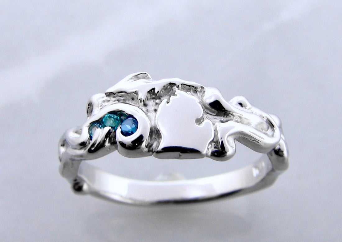 Silver ring with curls and waves surrounding a raised cutout shape of the state of Michigan. To the left of the Lower Peninsula are set three round blue gems. 