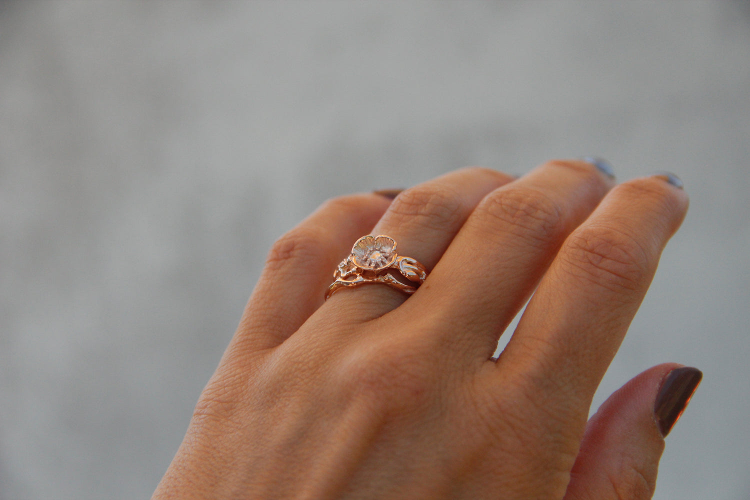 Image shows woman wearing bridal ring set with a sculpted poppy shape. The flower holds a solitaire diamond set low into the center in prongs shaped like poppy stamen. Polished rose gold show up against a gray backdrop