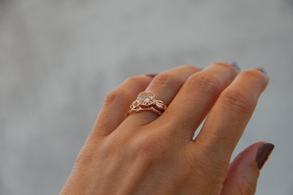 Image shows woman wearing bridal ring set with a sculpted poppy shape. The flower holds a solitaire diamond set low into the center in prongs shaped like poppy stamen. Polished rose gold show up against a gray backdrop