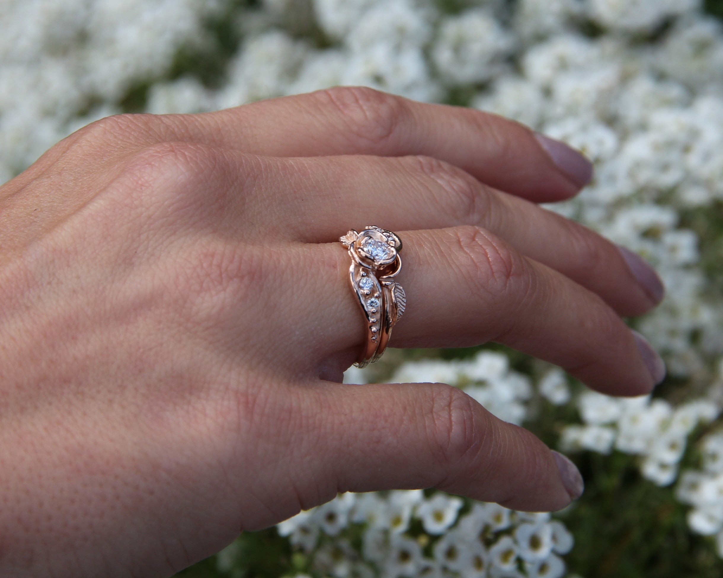 Photo shows a woman’s hand wearing a rose blooming on a rose gold wedding ring set that measures half an inch wide. The rose is 0.25 inch across and holds a round diamond, and the bands curve to fit together. Small raised beads are on each band