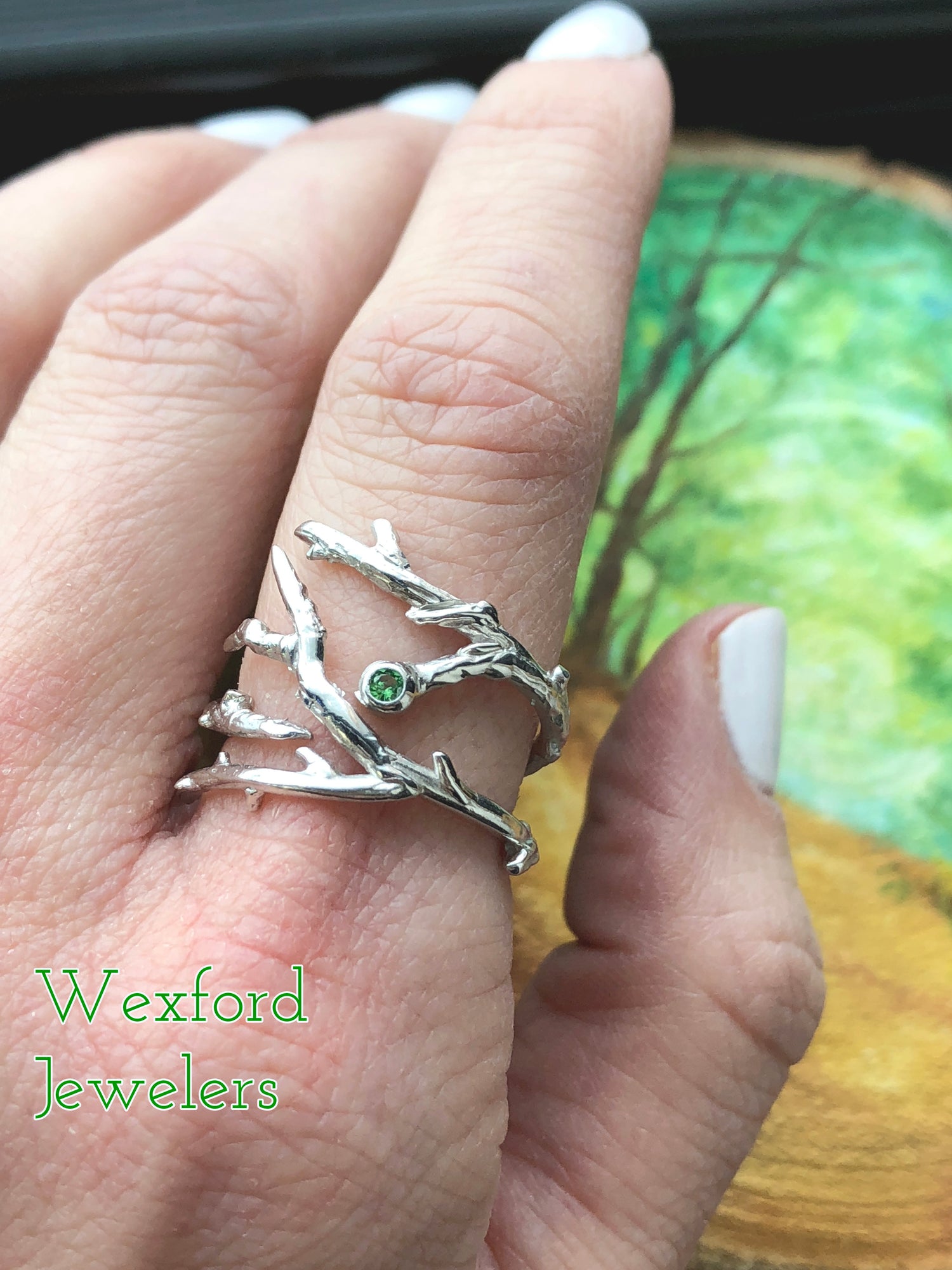 Woman’s hand wears a ring comprised of slender twigs wrapped around the finger. The branches span appx an inch on top, and in the center is a small green gem bezel set