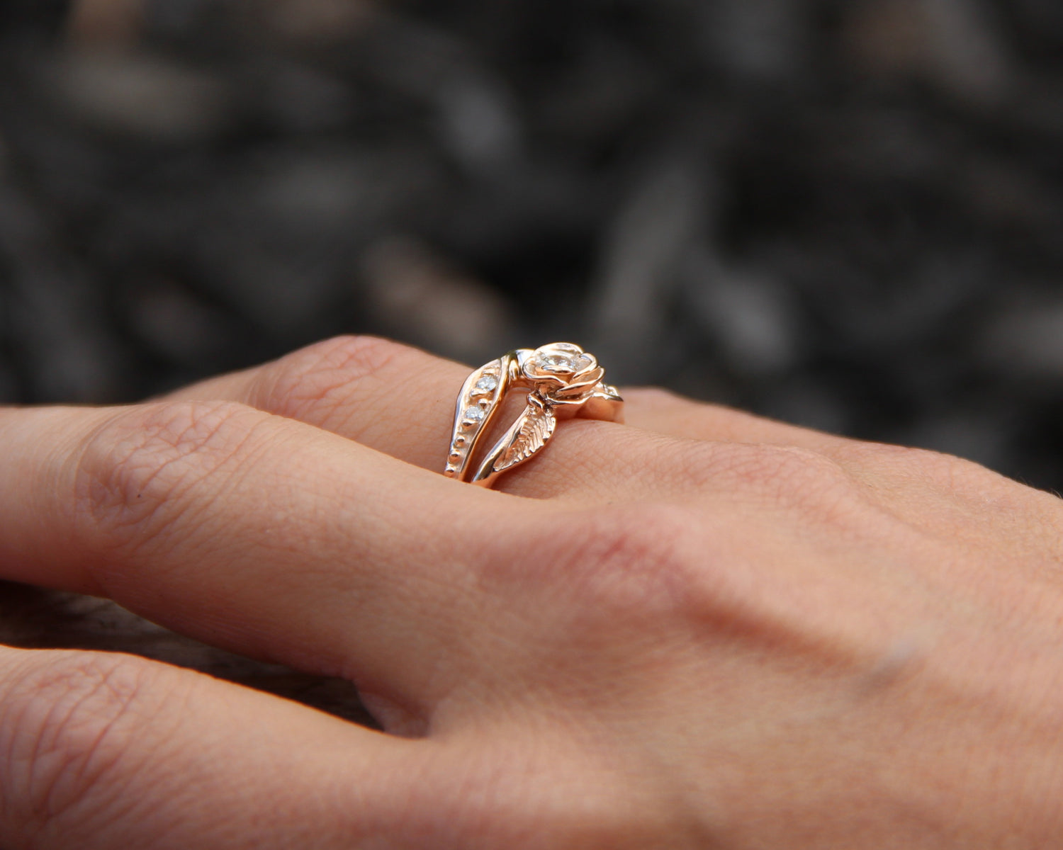  woman’s hand wears a rose bloom on a rose gold wedding ring set that measures half an inch wide. The rose is 0.25 inch across and holds a round diamond, and the bands curve to fit together. Small leaves and diamonds offset each side