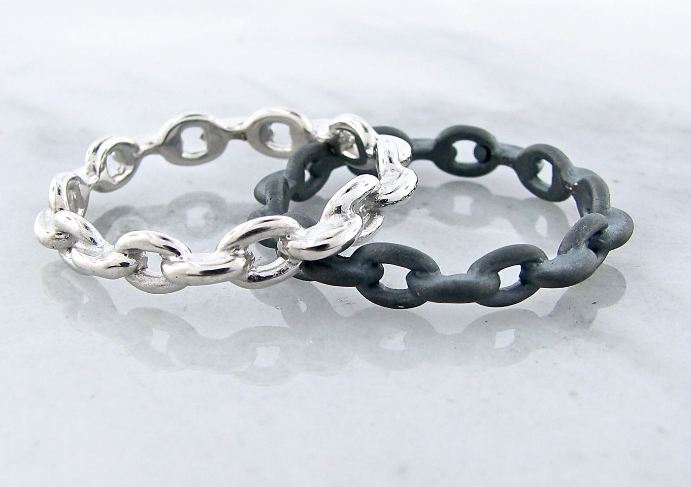 image shows two silver bands made of fused chain links. one band is high polished silver finish, the other is matte black
